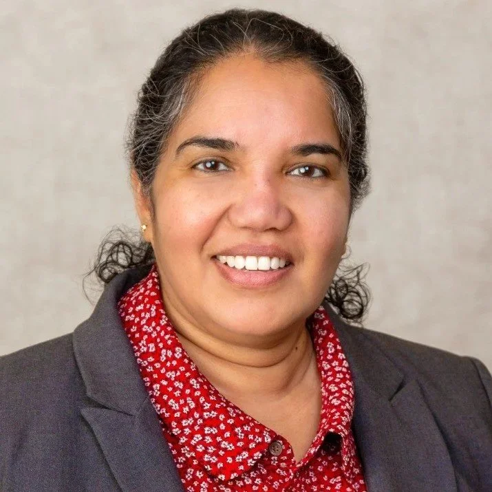 A woman with dark hair tied back, smiling, wearing a dark blazer and a red blouse with small white floral pattern, standing against a neutral background.