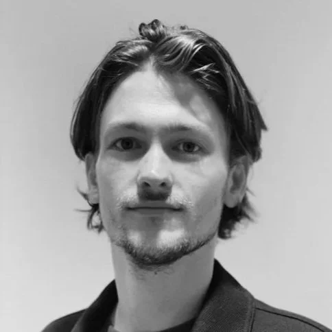 Black and white portrait of a young man with medium-length hair and light facial hair, wearing a dark jacket, against a plain background.