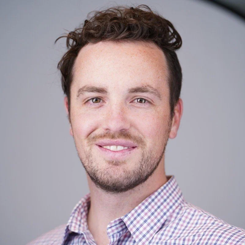 Portrait of a young man with brown curly hair, a beard, wearing a checkered shirt, smiling, against a neutral background.