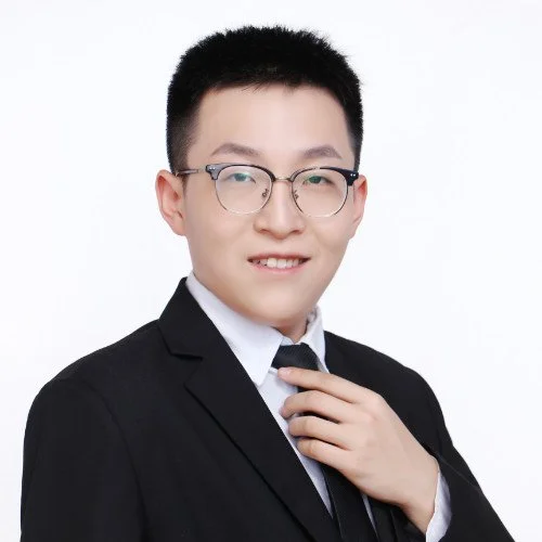A young man with short black hair, glasses, wearing a black suit, white shirt, and black tie, adjusting his tie and smiling for the camera.
