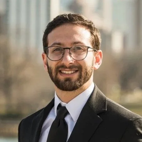 A man with glasses, a beard, and short hair, wearing a black suit and tie, smiling outdoors with buildings in the background.