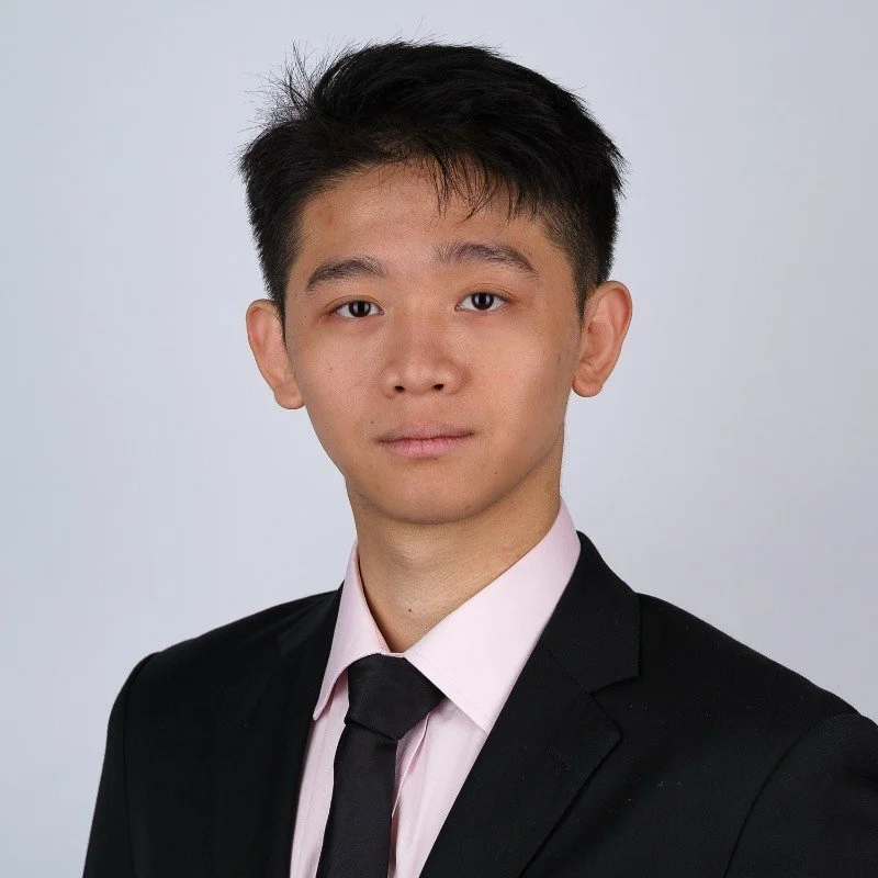 A young man with short black hair wearing a black suit, light pink shirt, and black tie, looking directly at the camera with a serious expression, against a plain gray background.