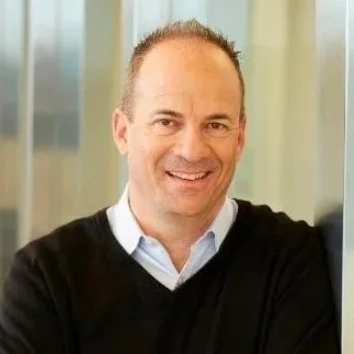 A smiling man with short hair, wearing a light blue collared shirt and black sweater, standing indoors with glass windows in the background.