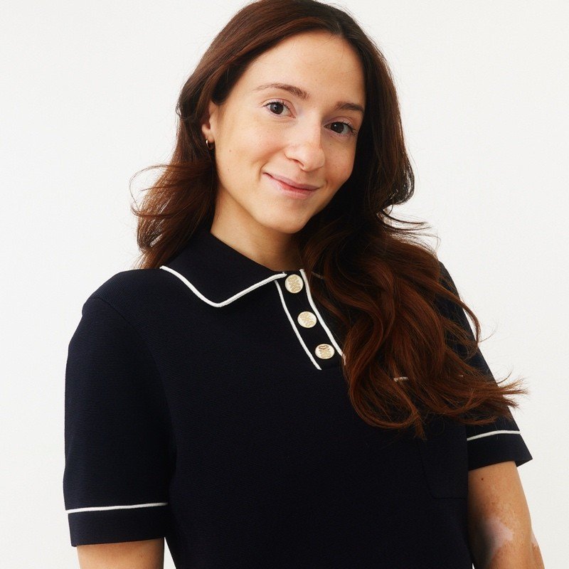 A young woman with long, wavy red hair and light skin, smiling slightly, wearing a black collared shirt with white piping and gold buttons, standing in front of a plain white background.