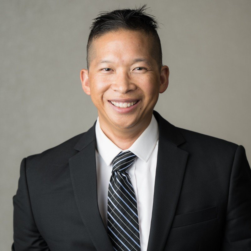 Portrait of a smiling man with short black hair, wearing a black suit, white shirt, and striped tie, against a plain background.