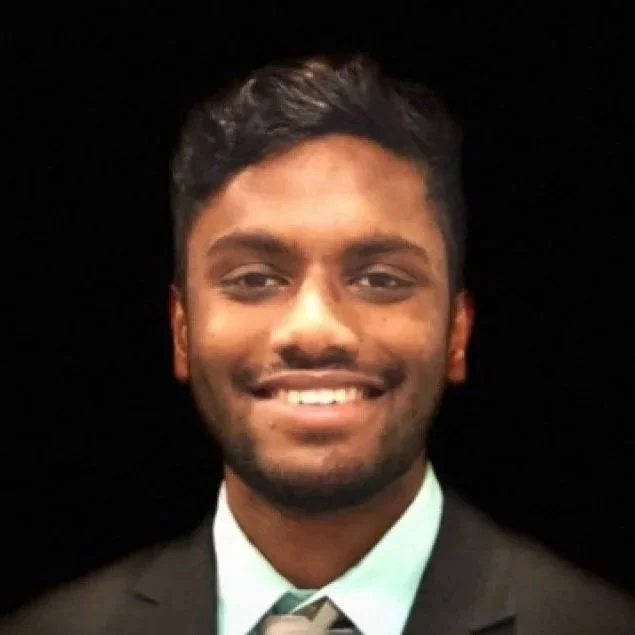 A young man with dark hair and a beard, wearing a suit and tie, smiling at the camera.