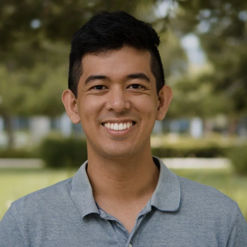 A young man with short black hair smiling outdoors, wearing a gray collared shirt with a park and trees blurred in the background.