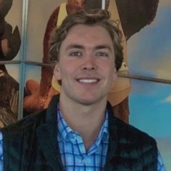 A smiling young man with light brown hair, wearing a blue checkered shirt and a dark jacket, standing indoors in front of a colorful background.
