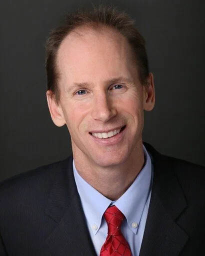 Portrait of a smiling man wearing a dark suit, light blue shirt, and red tie, against a dark background.