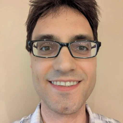Close-up of a man with short brown hair, glasses, and a light-colored shirt smiling at the camera.