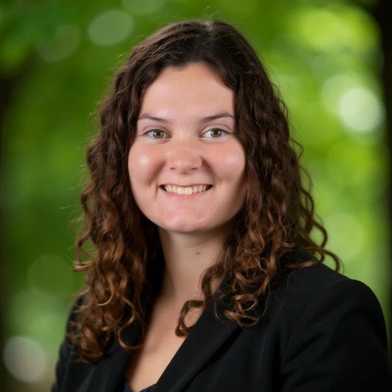 Portrait of a young woman with curly brown hair, smiling in front of a green outdoor background.
