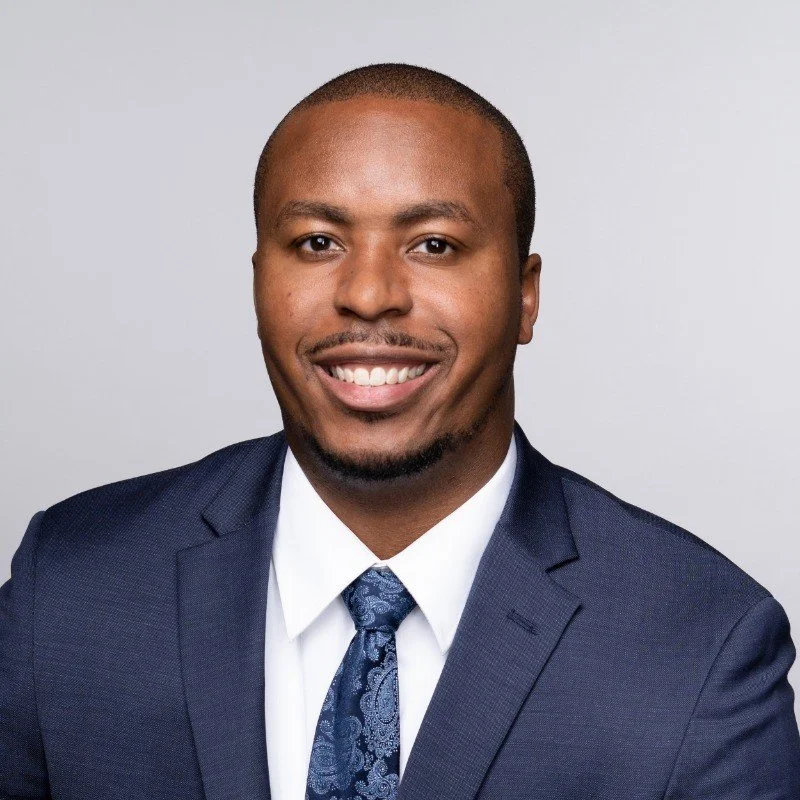 Headshot of a smiling African American man in a navy suit, white shirt, and patterned blue tie, against a plain light gray background.