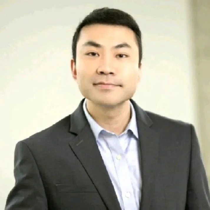 A young man with short dark hair wearing a dark gray suit jacket and a light blue collared shirt, standing against a beige wall.