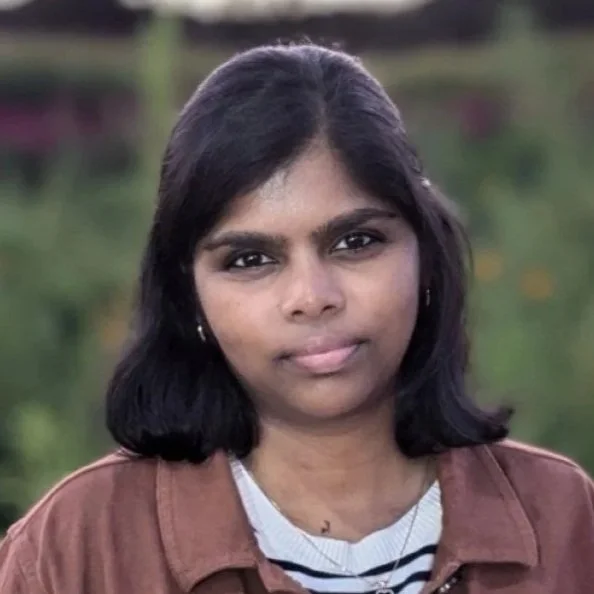 A woman with medium-length black hair, wearing a brown jacket over a striped shirt, stands outdoors with blurred green foliage in the background.