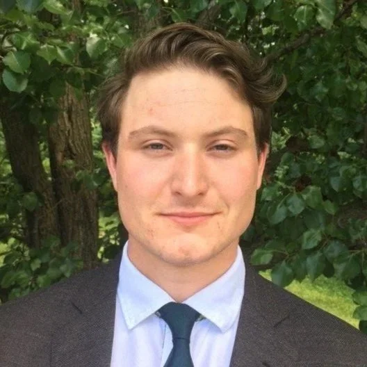 A young man in a suit and tie standing outdoors with green foliage in the background.