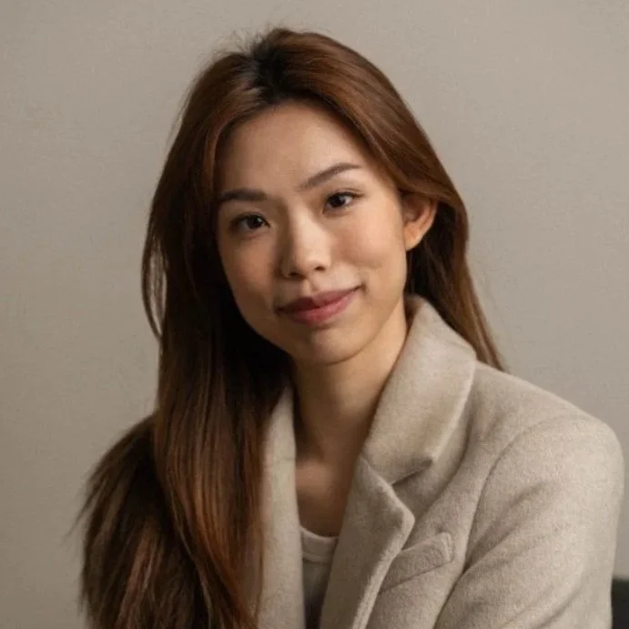 Young woman with long brown hair, smiling, wearing a light-colored coat, sitting indoors against a plain wall.