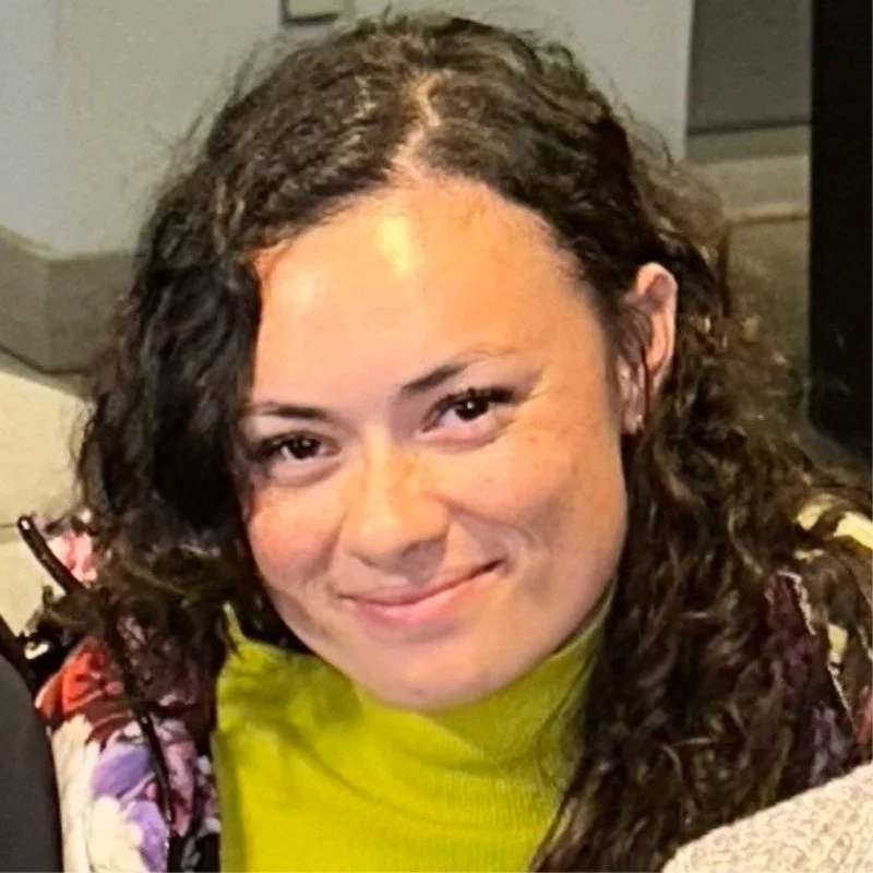 A woman with curly dark hair, wearing a bright yellow top, smiling at the camera.