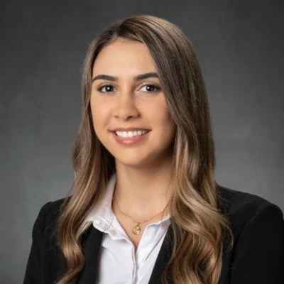 A young woman with long brown hair and a friendly smile, wearing a white shirt and black blazer, against a gray background.