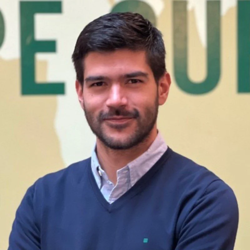 A man with dark hair and a beard in a collared shirt and navy sweater, standing against a yellow background with large green text.