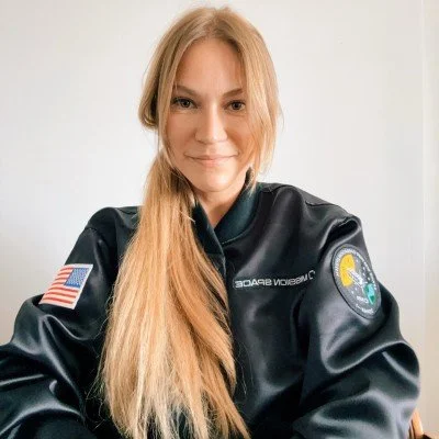 A woman with long blonde hair smiling, wearing a black NASA jacket with an American flag patch and a NASA logo patch on the sleeves.