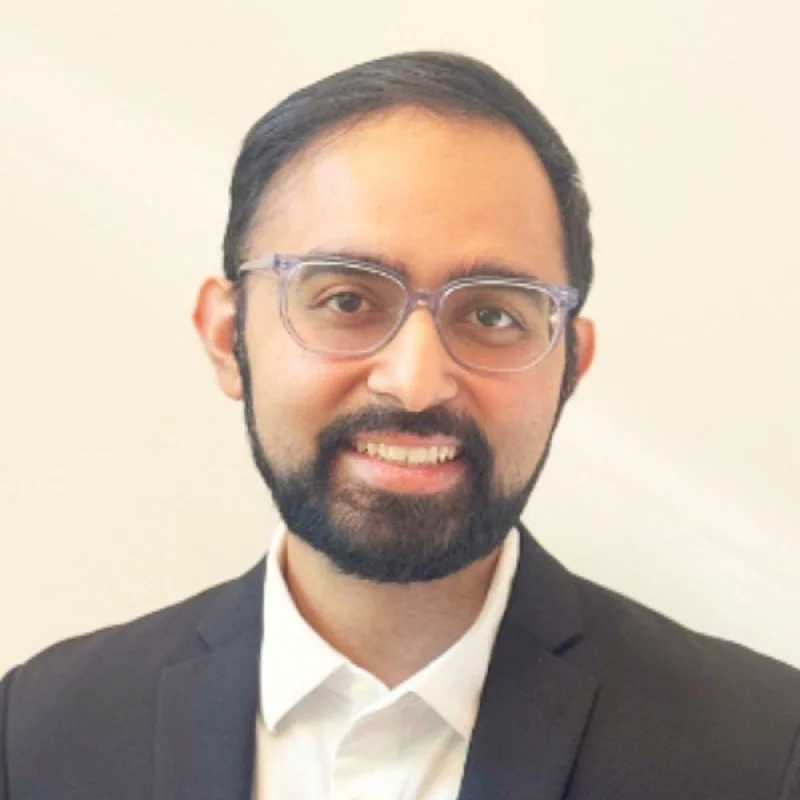 A man with dark hair, glasses, and a beard smiling in front of a plain light-colored background, dressed in a black suit and white shirt.