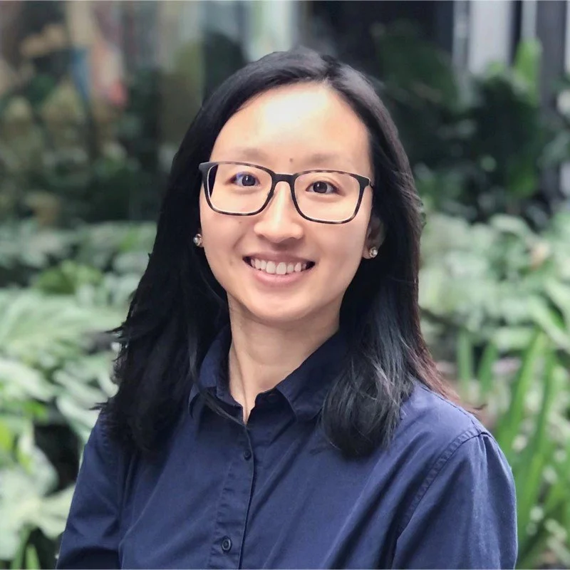 A woman with black hair and glasses smiling at the camera, wearing a dark blue shirt, with green plants in the background.