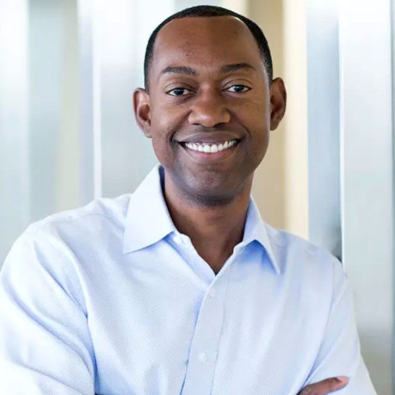 A smiling man in a light blue dress shirt standing indoors near glass windows.