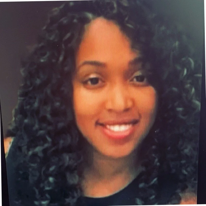 Close-up of a smiling woman with dark, curly hair and medium skin tone.