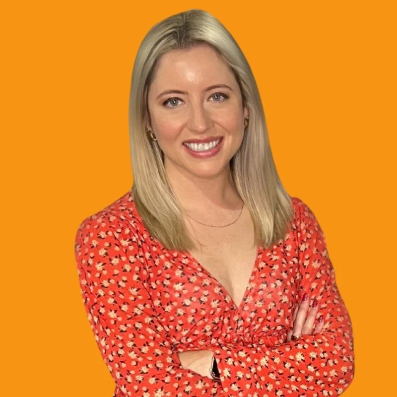 A woman with blonde hair smiling, wearing a red floral blouse, standing with arms crossed against an orange background.