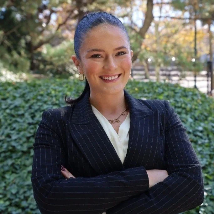 A woman with dark hair in a ponytail, wearing a black pinstripe blazer over a white top, smiling and crossing her arms outdoors with green bushes and trees in the background.