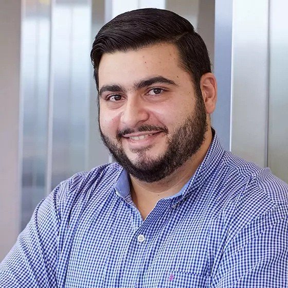 Portrait of a man with dark hair and beard wearing a blue checkered shirt, smiling, indoor setting with windows in the background.