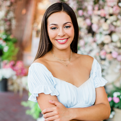 Young woman with long dark hair smiling outdoors, surrounded by pink and white flowers.