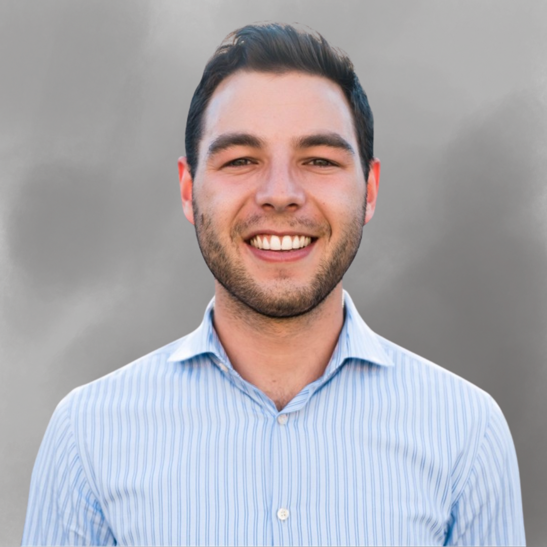 A smiling young man with short dark hair and a beard, wearing a light blue striped button-up shirt, standing outdoors on a cloudy day.