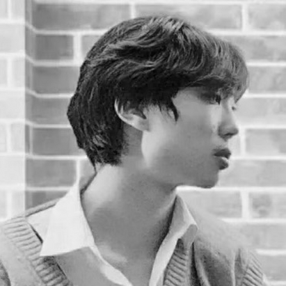 Profile of a young man with dark, medium-length hair, wearing a collared shirt and sweater, in front of a brick wall.