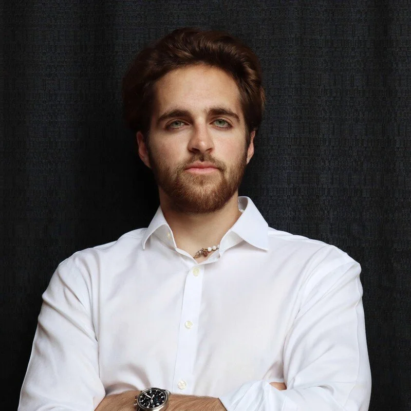 A young man with brown hair and a beard wearing a white dress shirt, standing against a dark textured background.