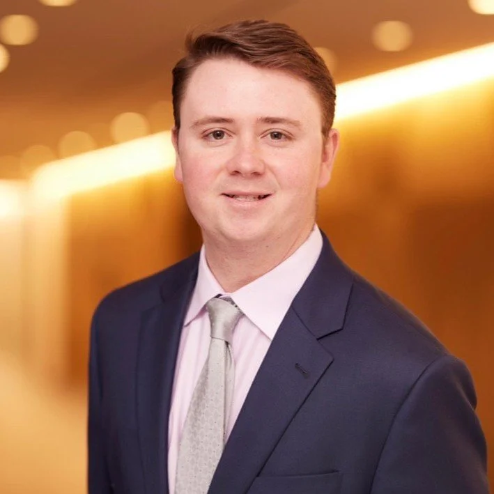 Professional headshot of a young man in a navy suit, white shirt, and light-colored tie, standing in front of a blurred warm-toned background.