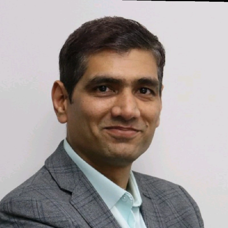A professional man with dark hair, wearing a gray checkered suit jacket and a light blue shirt, standing in front of a plain white wall and smiling at the camera.