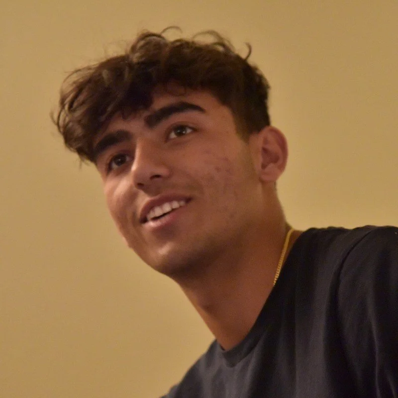 A young man with curly brown hair and a slight smile, wearing a black shirt, looking slightly to the side against a plain beige background.