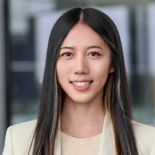 Portrait of a young Asian woman with long black hair and a slight smile, wearing a cream blazer and sweater, with a blurred background of office buildings.