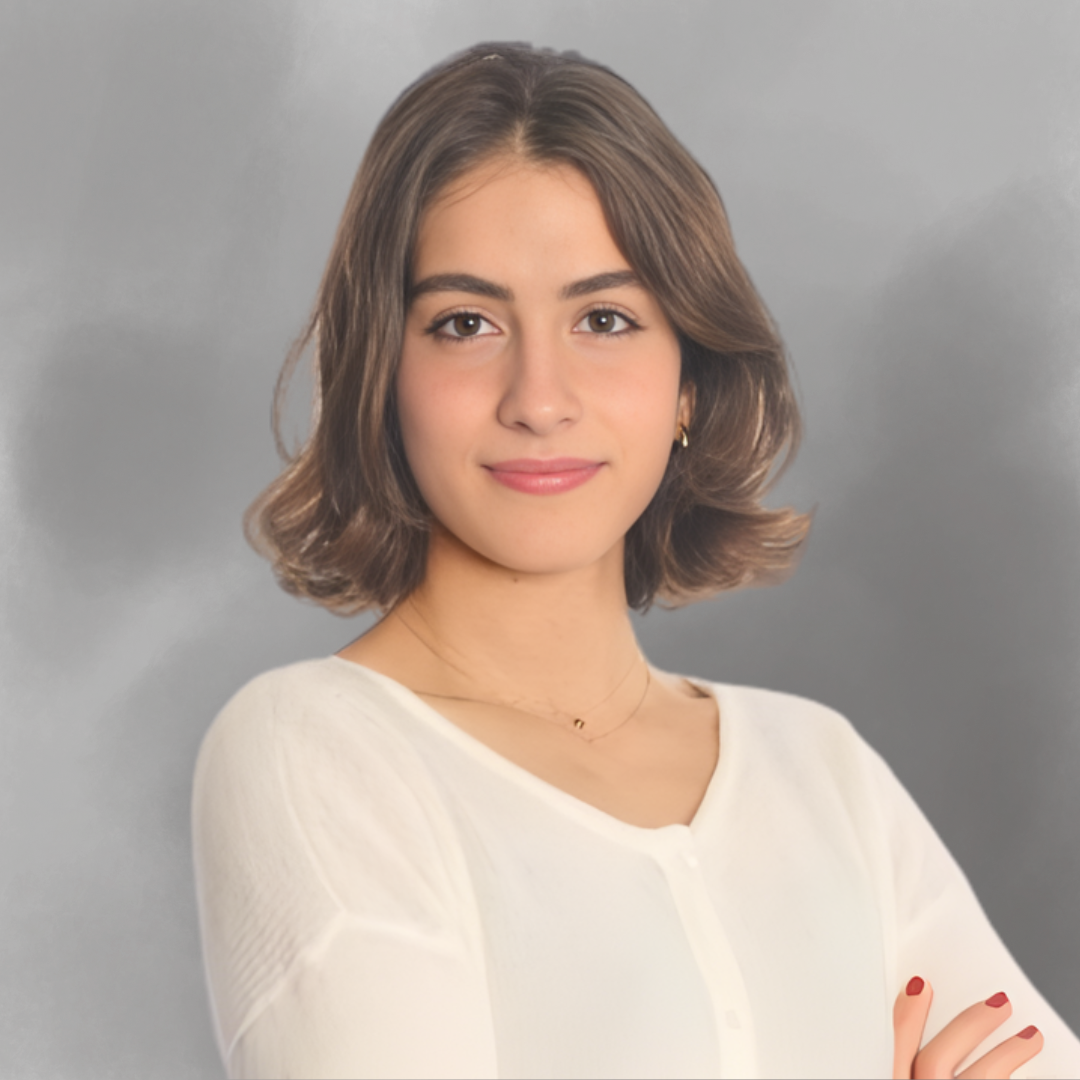 Portrait of a young woman with shoulder-length brown hair, wearing a white top, gold earrings, and a delicate necklace, posed with arms crossed against a gray background.