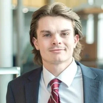 A young man with medium-length brown hair, wearing a dark suit, white shirt, and striped red tie, smiling confidently in a professional setting.
