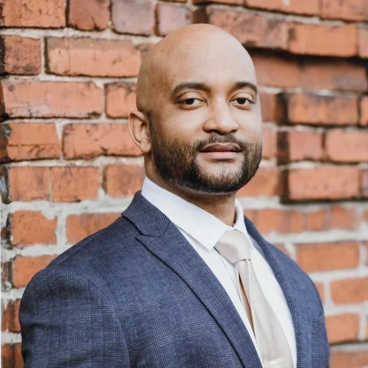 A man wearing a dark blue suit and a beige tie, standing against a red brick wall.