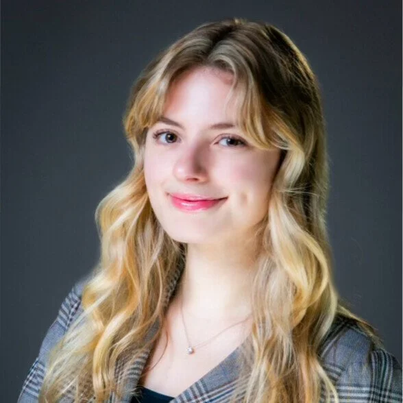 Portrait of a young woman with long blonde hair, smiling, wearing a checkered blazer and a necklace, against a dark gray background.