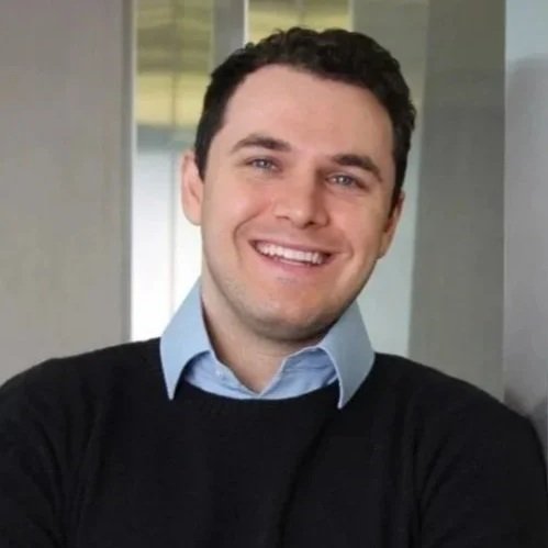 A smiling man with short dark hair, wearing a light blue collared shirt and a black sweater, standing indoors.