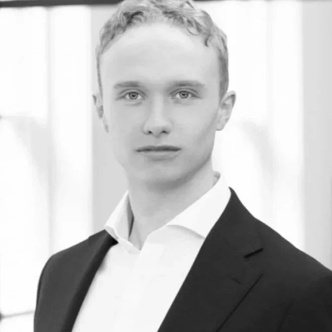 Portrait of a young man in a suit and white shirt, looking at the camera with a serious expression, in a bright indoor setting.