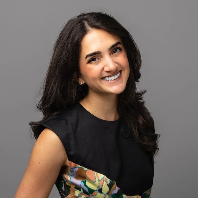 A woman with long dark hair smiling, wearing a black top with a colorful abstract pattern on the front, standing against a gray background.