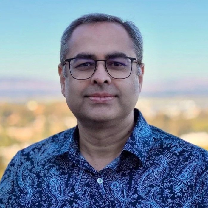 A man with glasses and short gray hair wearing a blue patterned button-up shirt, outdoors with a blurred landscape and sky in the background.