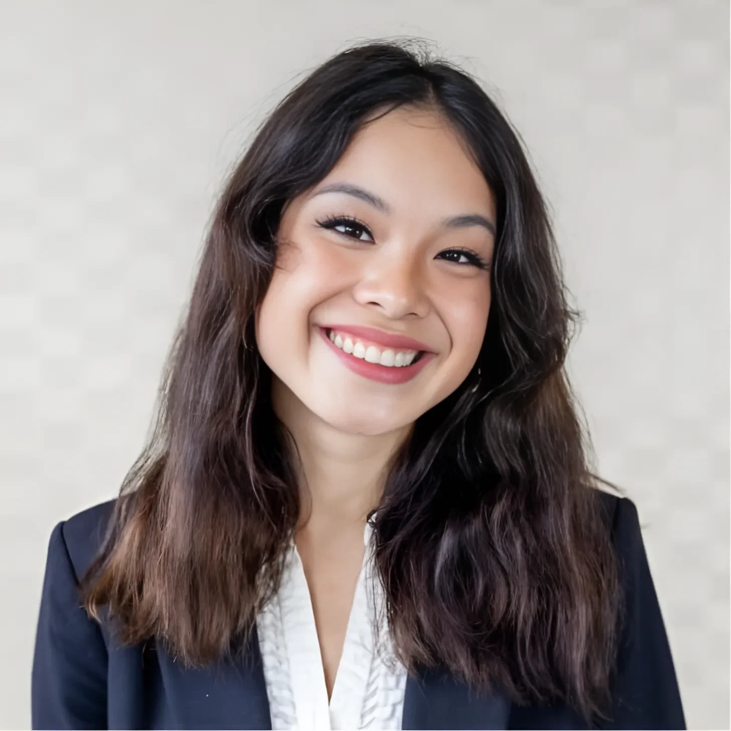 A young woman with long wavy dark hair, smiling, wearing a black blazer and white blouse, against a light neutral background.