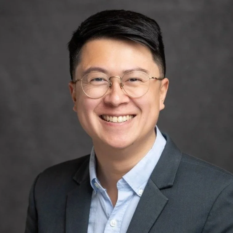 A young man with glasses and short black hair, smiling, wearing a dark suit and light blue dress shirt against a dark gray background.