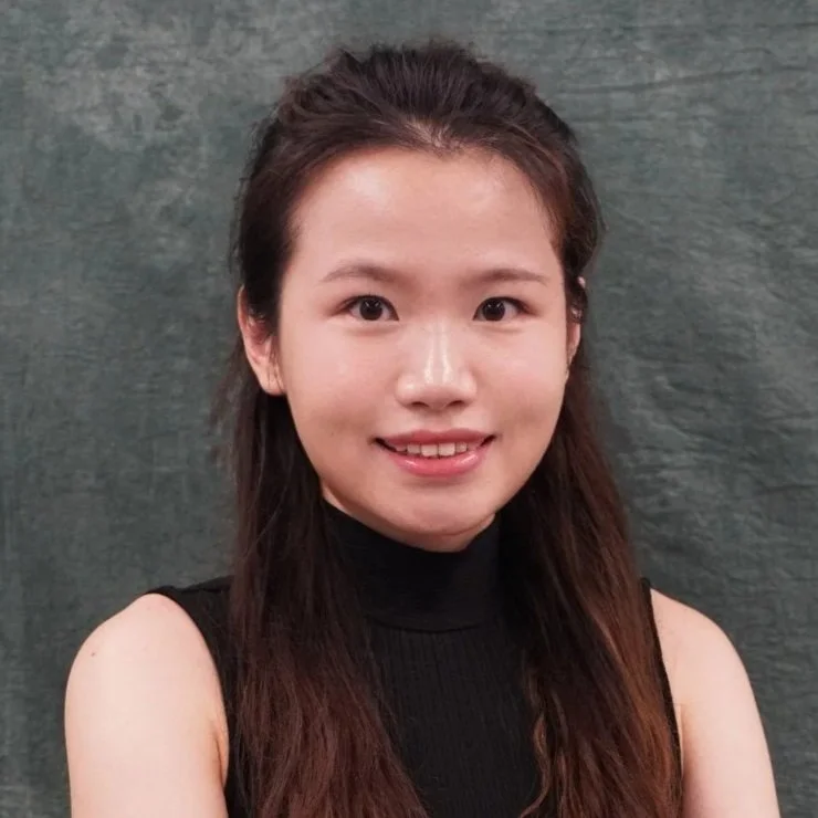 A young woman with long brown hair, wearing a black sleeveless top, smiling in front of a grey textured background.